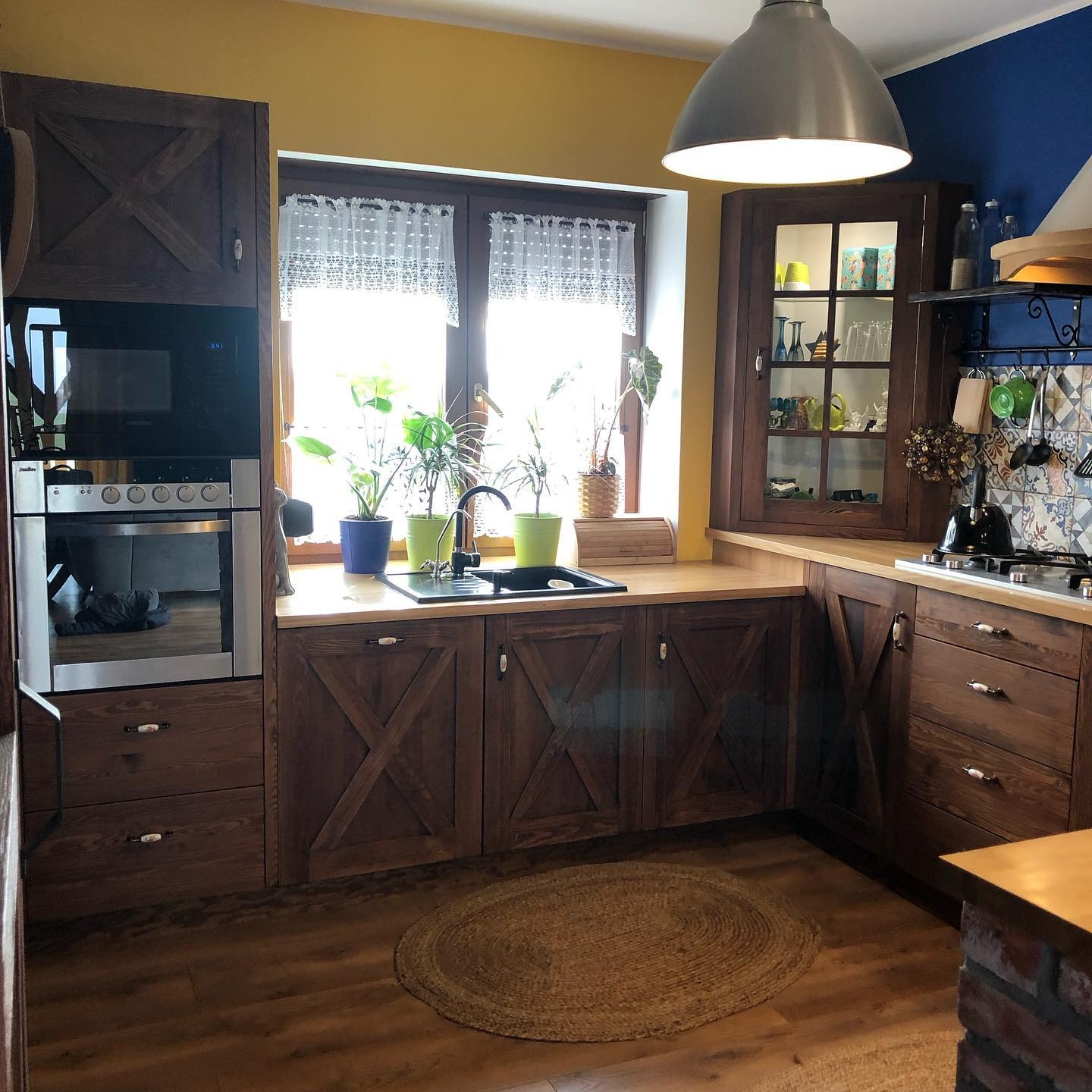 Rustykalna kuchnia z drewnianymi frontami szafek w kształcie krzyża, okrągłym dywanikiem na podłodze i roślinami w doniczkach na parapecie.