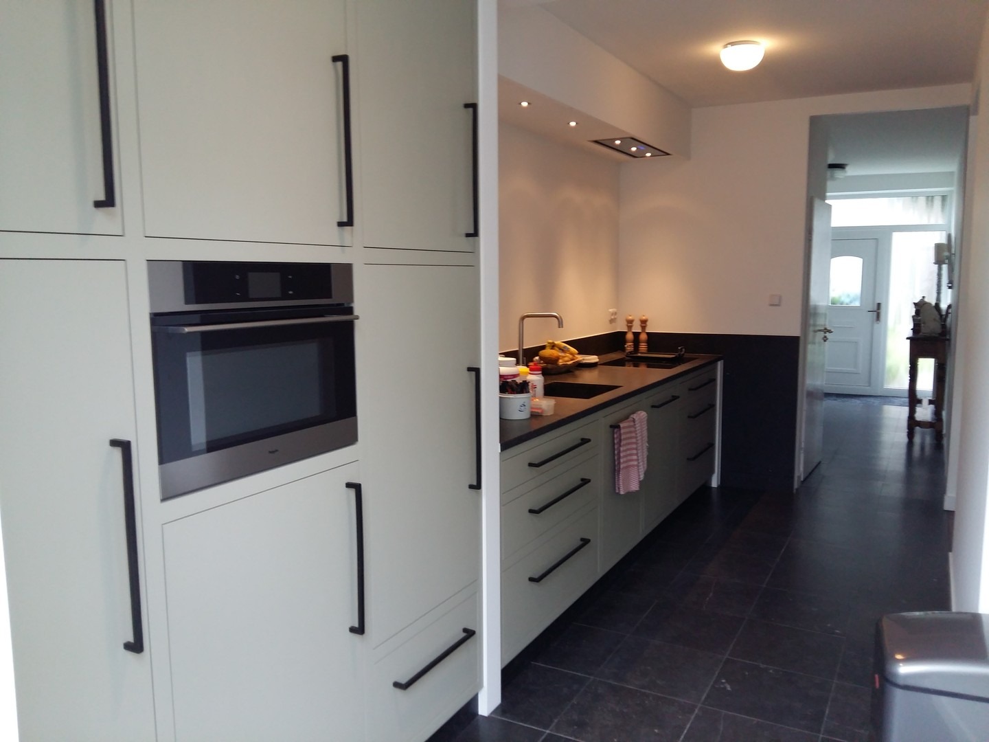 Nowoczesna kuchnia z jasnymi szafkami z długimi, czarnymi uchwytami i wbudowanym piekarnikiem, z ciemnym blatem i czarną ścianą w tle, widok na przedpokój z białymi drzwiami wejściowymi.