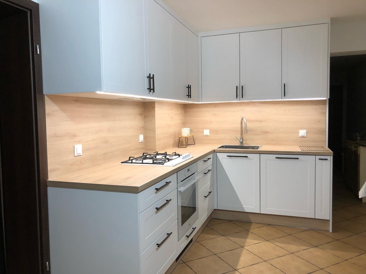 Jasna kuchnia z białymi szafkami i drewnianym blatem, oświetlenie LED pod szafkami wiszącymi, czarne, minimalistyczne uchwyty, płyta gazowa na blacie.