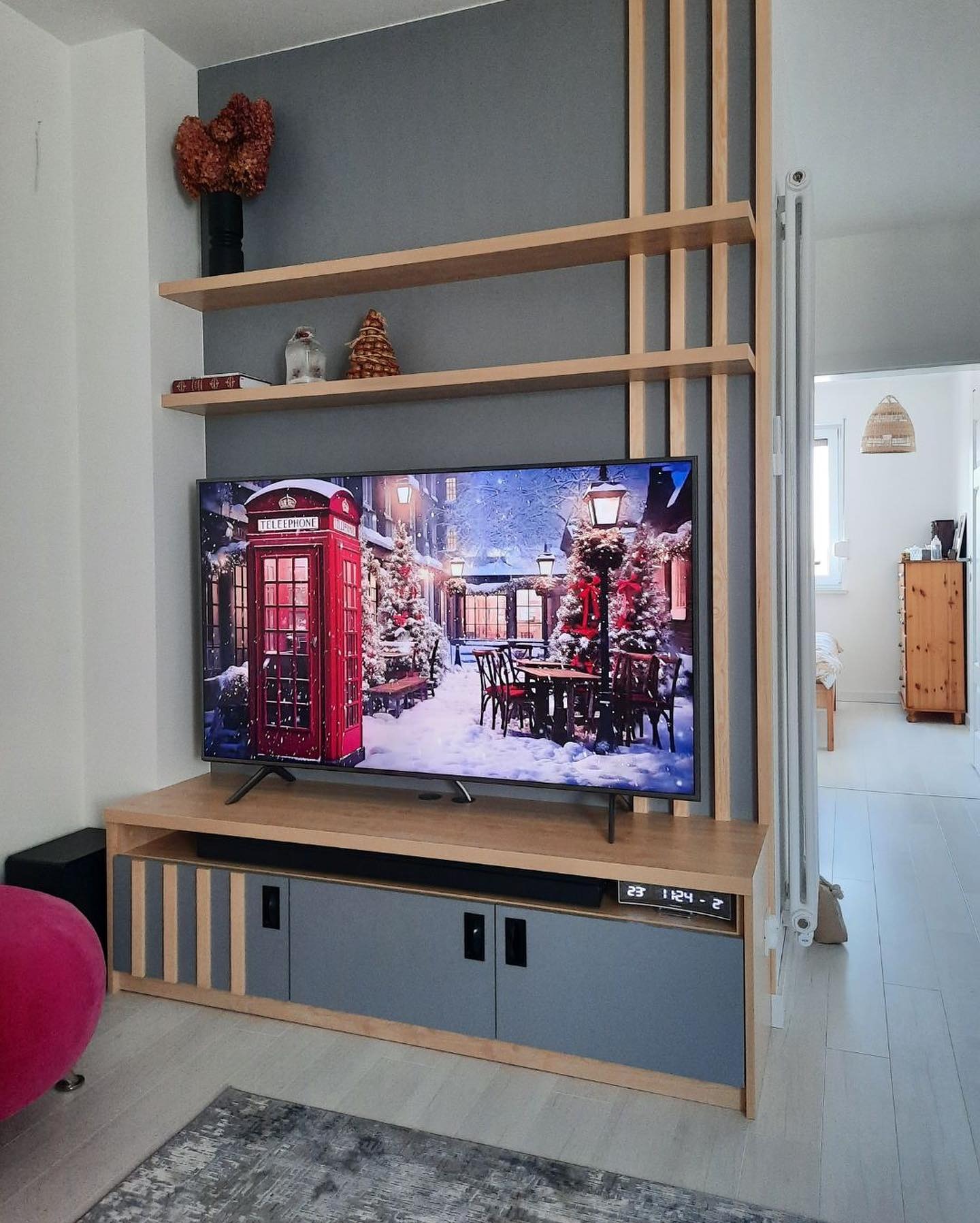 Nowoczesna szafka RTV w odcieniach drewna i szarości, z widocznymi elementami dekoracyjnymi i półkami, na tle szarej ściany z drewnianymi listwami.