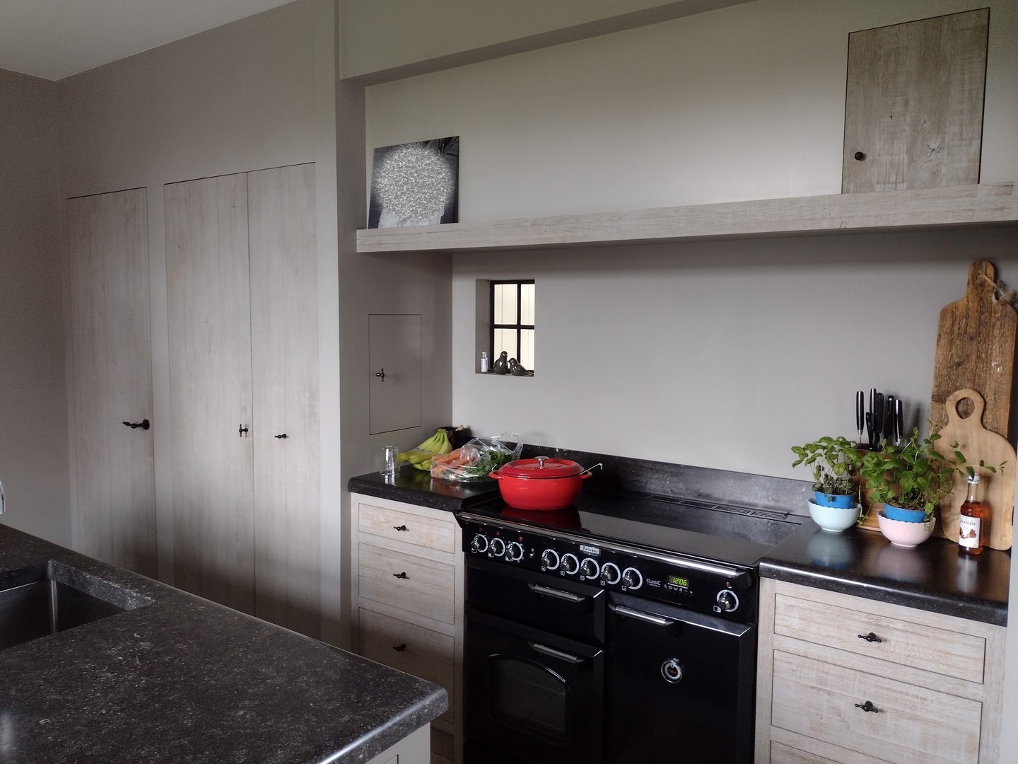 Jasna kuchnia z czarnym piecem wolnostojącym, blatem z ciemnego kamienia i drewnianymi frontami szafek z czarnymi uchwytami. Na blacie stoi czerwony garnek, deska do krojenia i zioła w doniczkach.