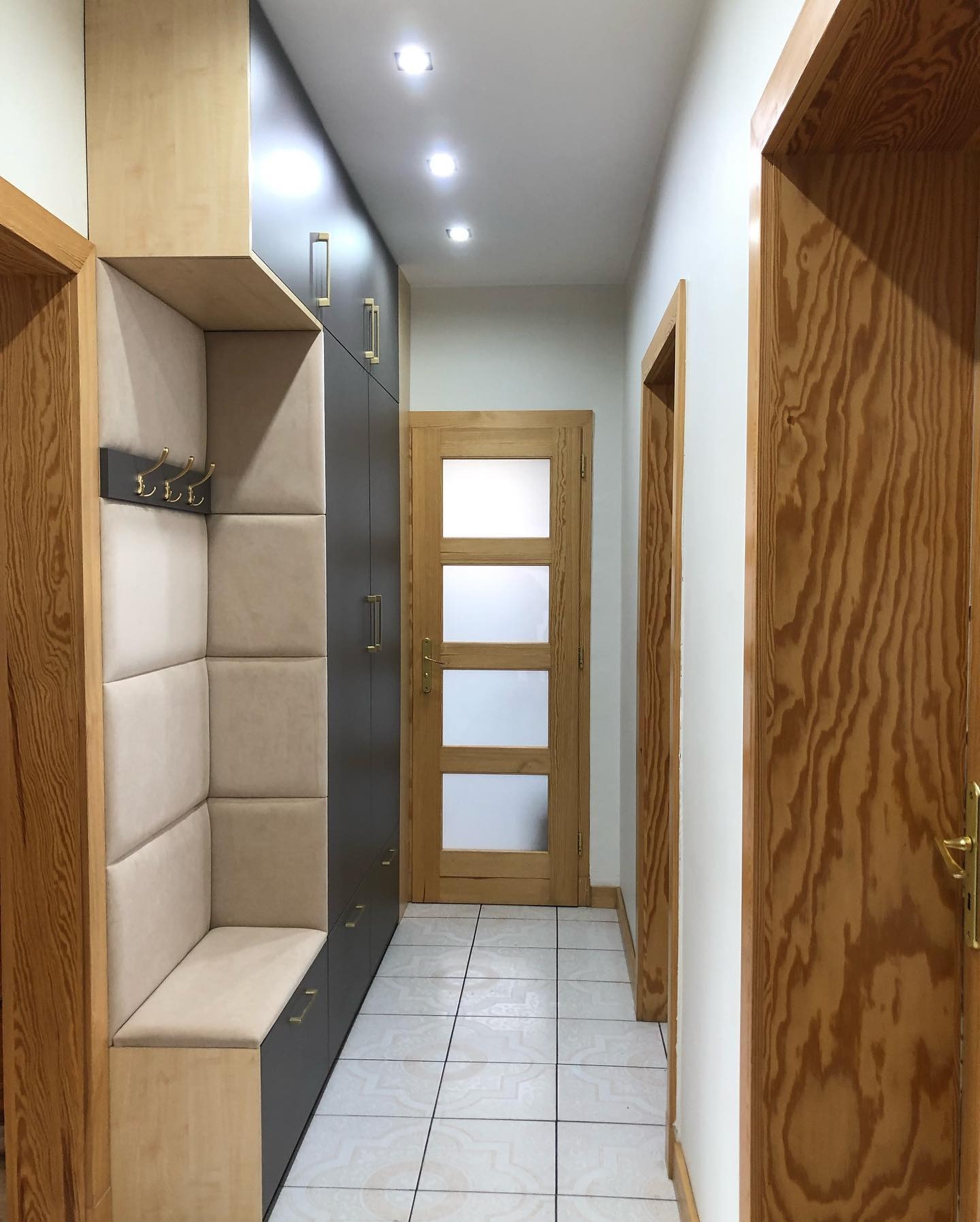 Wnętrze przedpokoju z zabudową meblową: szafa w kolorze grafitowym z podłużnymi, złotymi uchwytami, obok siedzisko z tapicerowanym oparciem i wieszakami na ścianie. W tle widoczne drewniane drzwi...