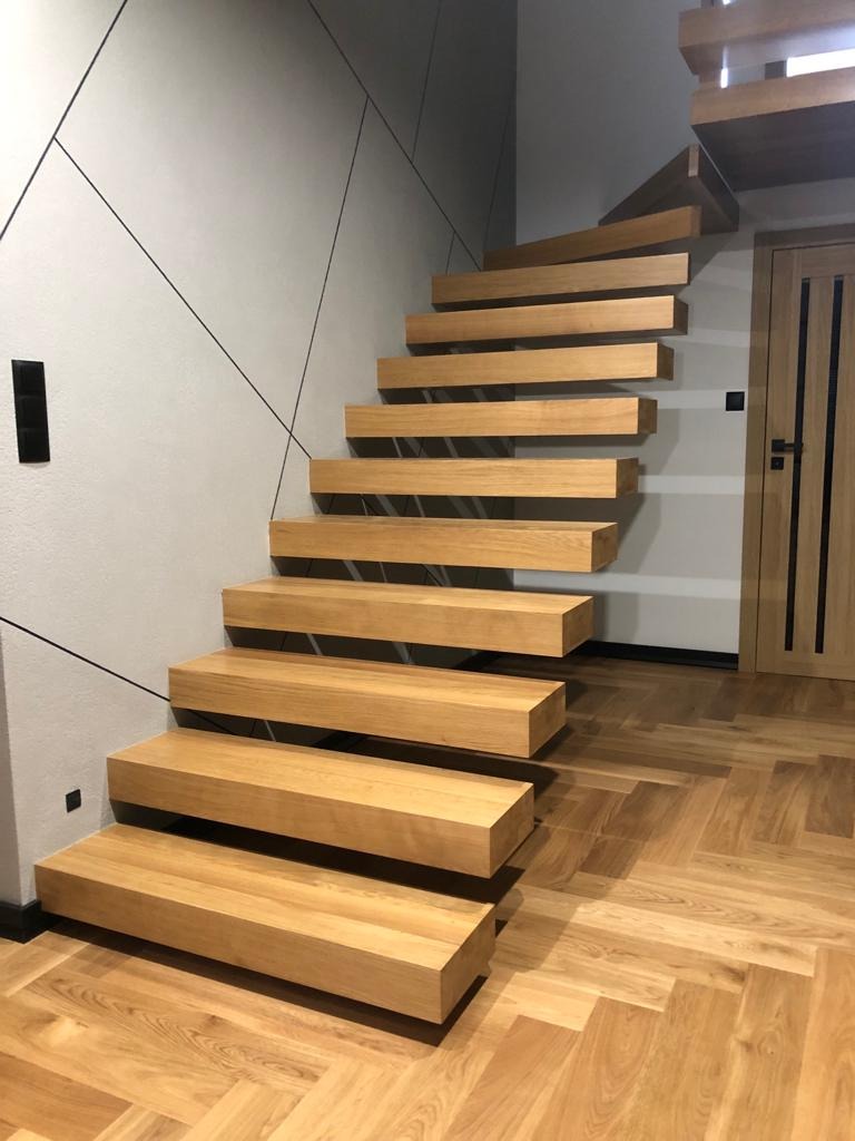 Nowoczesne, drewniane schody wspornikowe z jasnego drewna, zamontowane przy ścianie z geometrycznym wzorem, parkiet ułożony w jodełkę.