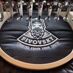 drukenhaft - Proces haftowania logo 'PIVOVSKY Crafted from your taste' na czarnej kurtce za pomocą maszyny hafciarskiej z widocznymi igłami i kolorowymi nićmi.