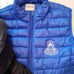 drukenhaft - Niebieski, pikowany bezrękawnik z logo 'Pivovsky' haftowanym na piersi, widoczny fragment czarnej kurtki obok.