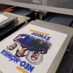 drukenhaft - Biała koszulka z nadrukiem kabaretu Neo-Nówka w maszynie drukarskiej, widoczne dysze drukujące i elementy mechaniczne urządzenia.