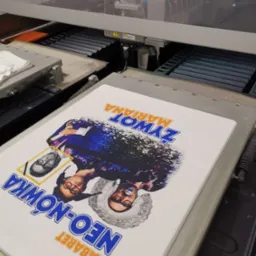 Biała koszulka z nadrukiem kabaretu Neo-Nówka w maszynie drukarskiej, widoczne dysze drukujące i elementy mechaniczne urządzenia.