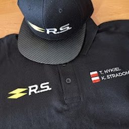drukenhaft - Czarna koszulka polo z wyhaftowanym logo R.S. i imionami oraz nazwiskami T. Nykiel i K. Stradomski wraz z flagą Austrii, leżąca obok czarnej czapki z daszkiem z logo R.S. na drewnianym stole.