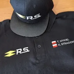Czarna koszulka polo z wyhaftowanym logo R.S. i imionami oraz nazwiskami T. Nykiel i K. Stradomski wraz z flagą Austrii, leżąca obok czarnej czapki z daszkiem z logo R.S. na drewnianym stole.