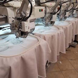 drukenhaft - Rząd automatycznych maszyn do szycia, wykonujących identyczny wzór na białych tkaninach, naciągniętych na okrągłe tamborki. Widoczny fragment osoby obsługującej maszyny.