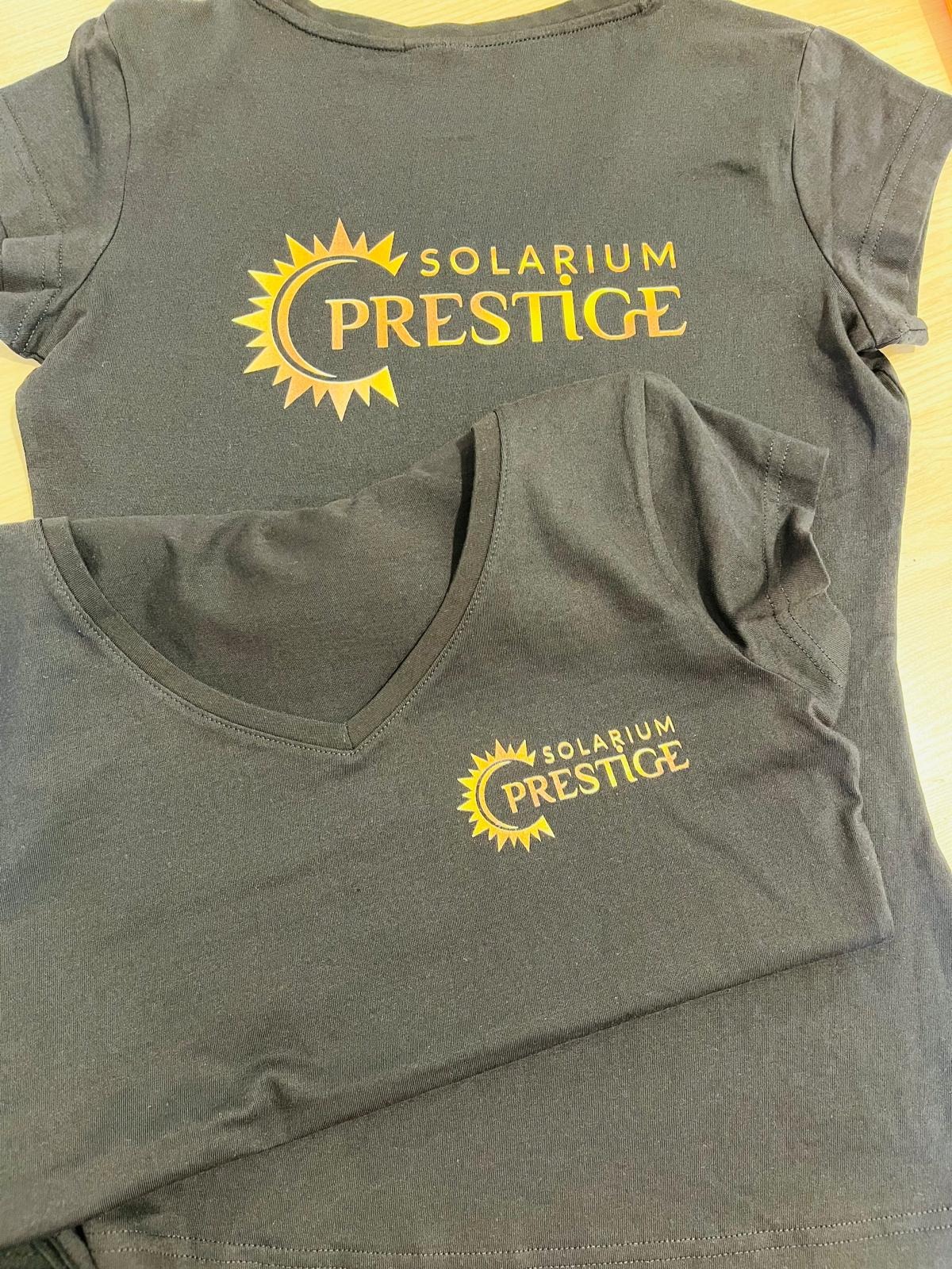 Dwie czarne koszulki, jedna z okrągłym dekoltem, druga z dekoltem w serek, obie z nadrukiem w kolorze złota przedstawiającym logo 'Solarium Prestige'.