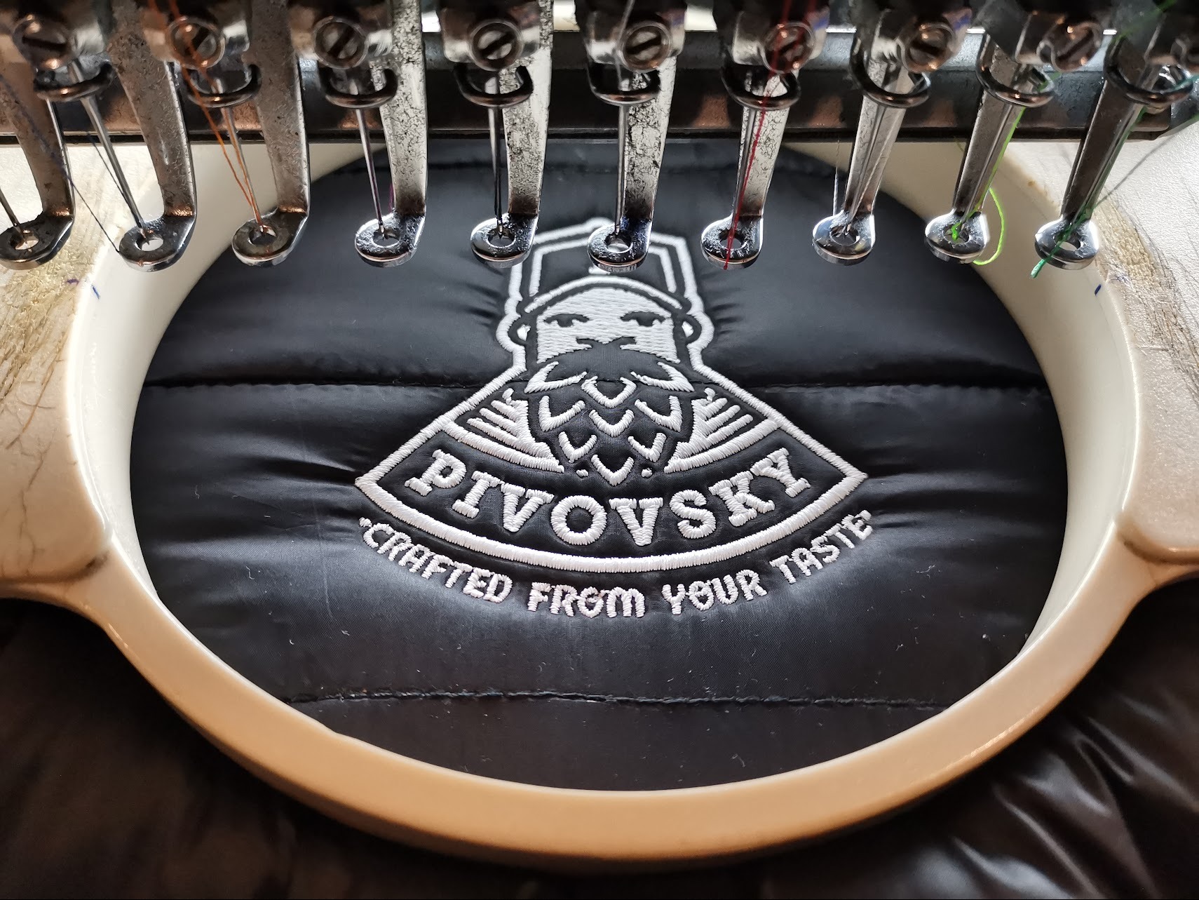 Proces haftowania logo 'PIVOVSKY Crafted from your taste' na czarnej kurtce za pomocą maszyny hafciarskiej z widocznymi igłami i kolorowymi nićmi.
