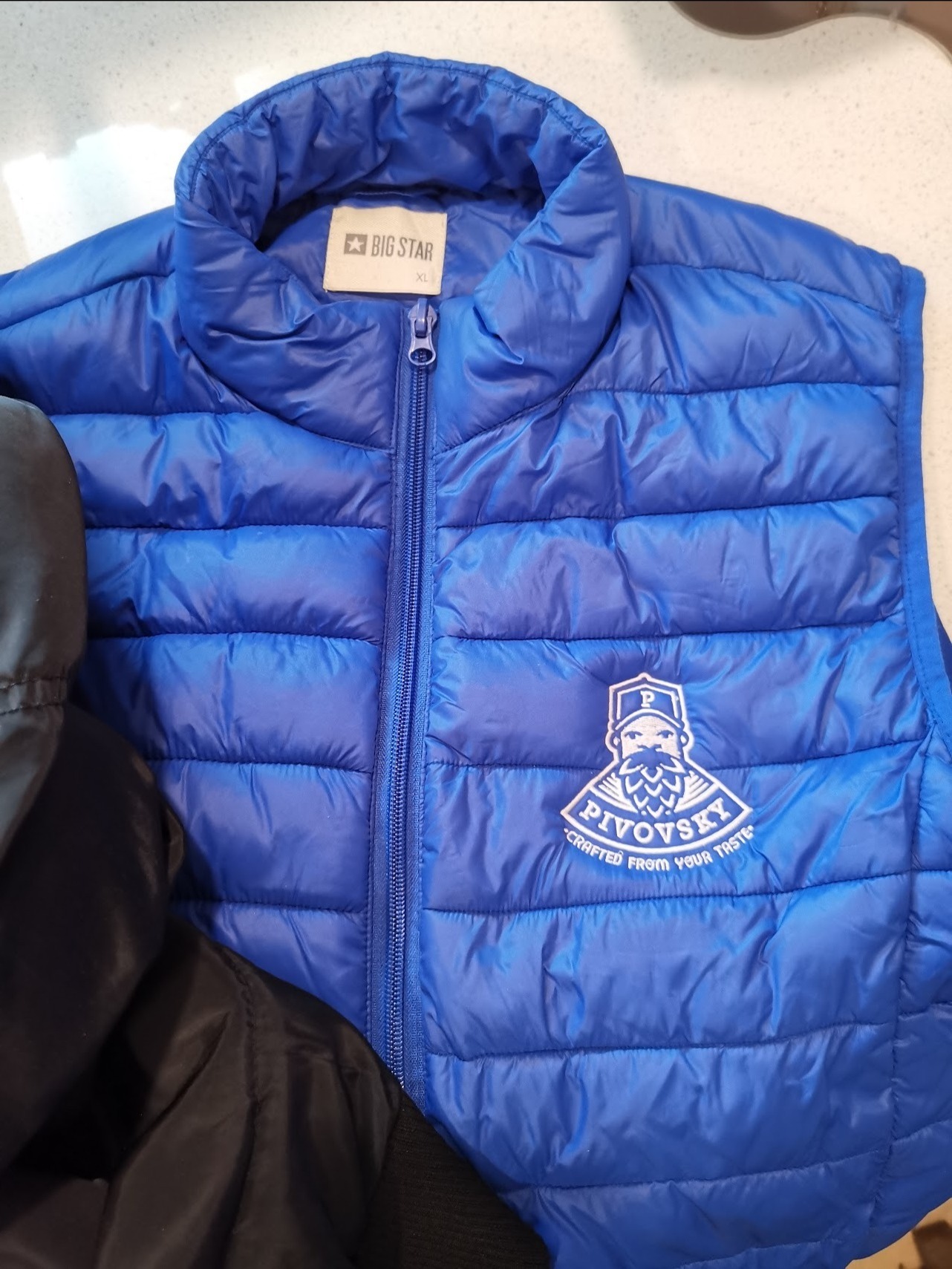Niebieski, pikowany bezrękawnik z logo 'Pivovsky' haftowanym na piersi, widoczny fragment czarnej kurtki obok.
