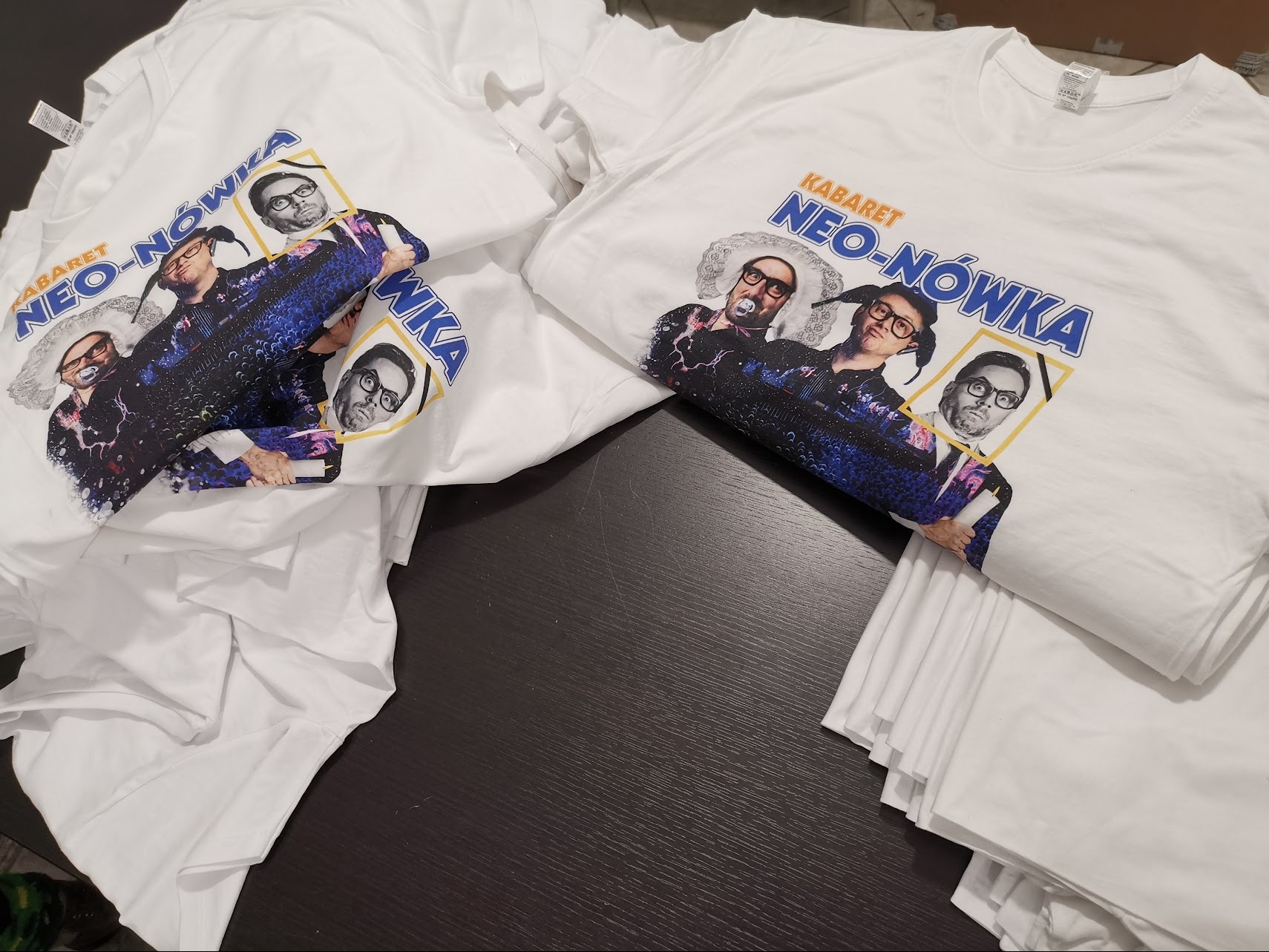 Białe koszulki z nadrukiem kabaretu Neo-Nówka ułożone na ciemnym blacie.