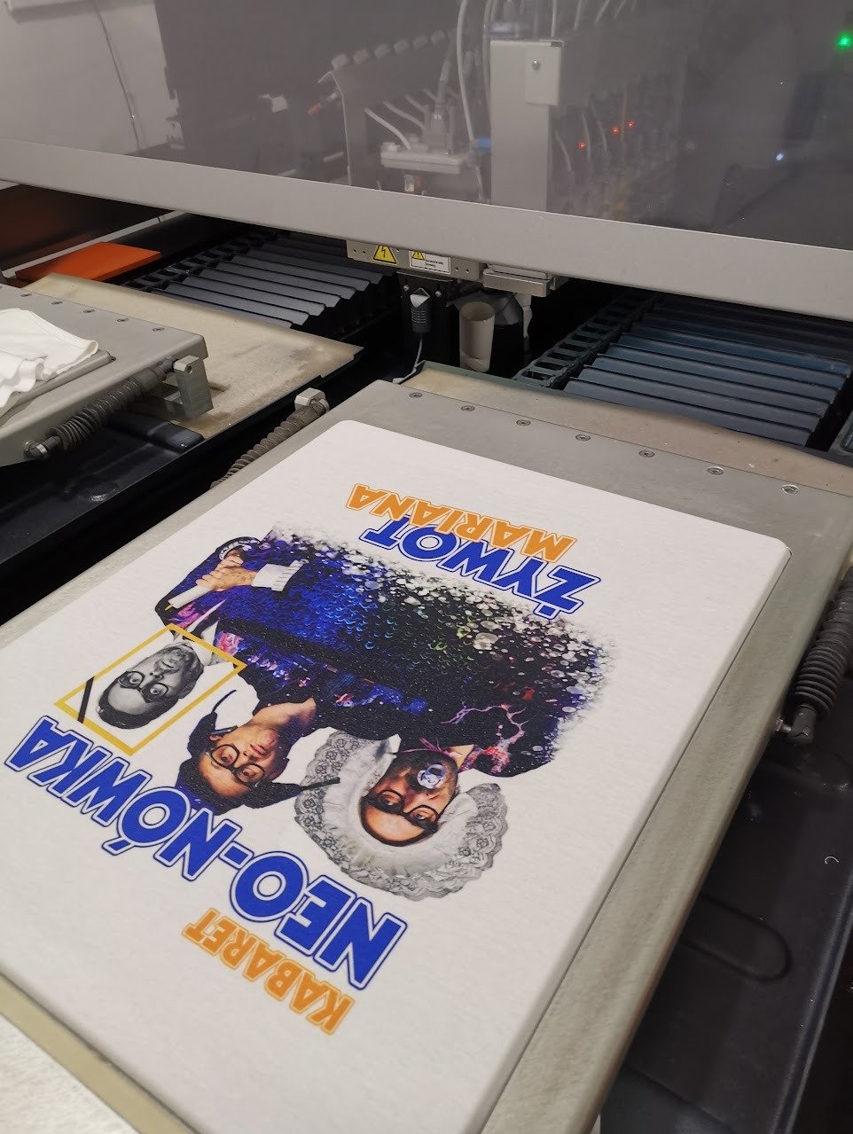 Biała koszulka z nadrukiem kabaretu Neo-Nówka w maszynie drukarskiej, widoczne dysze drukujące i elementy mechaniczne urządzenia.