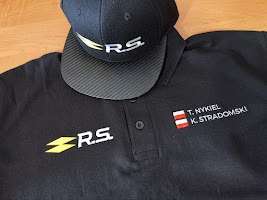 Czarna koszulka polo z wyhaftowanym logo R.S. i imionami oraz nazwiskami T. Nykiel i K. Stradomski wraz z flagą Austrii, leżąca obok czarnej czapki z daszkiem z logo R.S. na drewnianym stole.