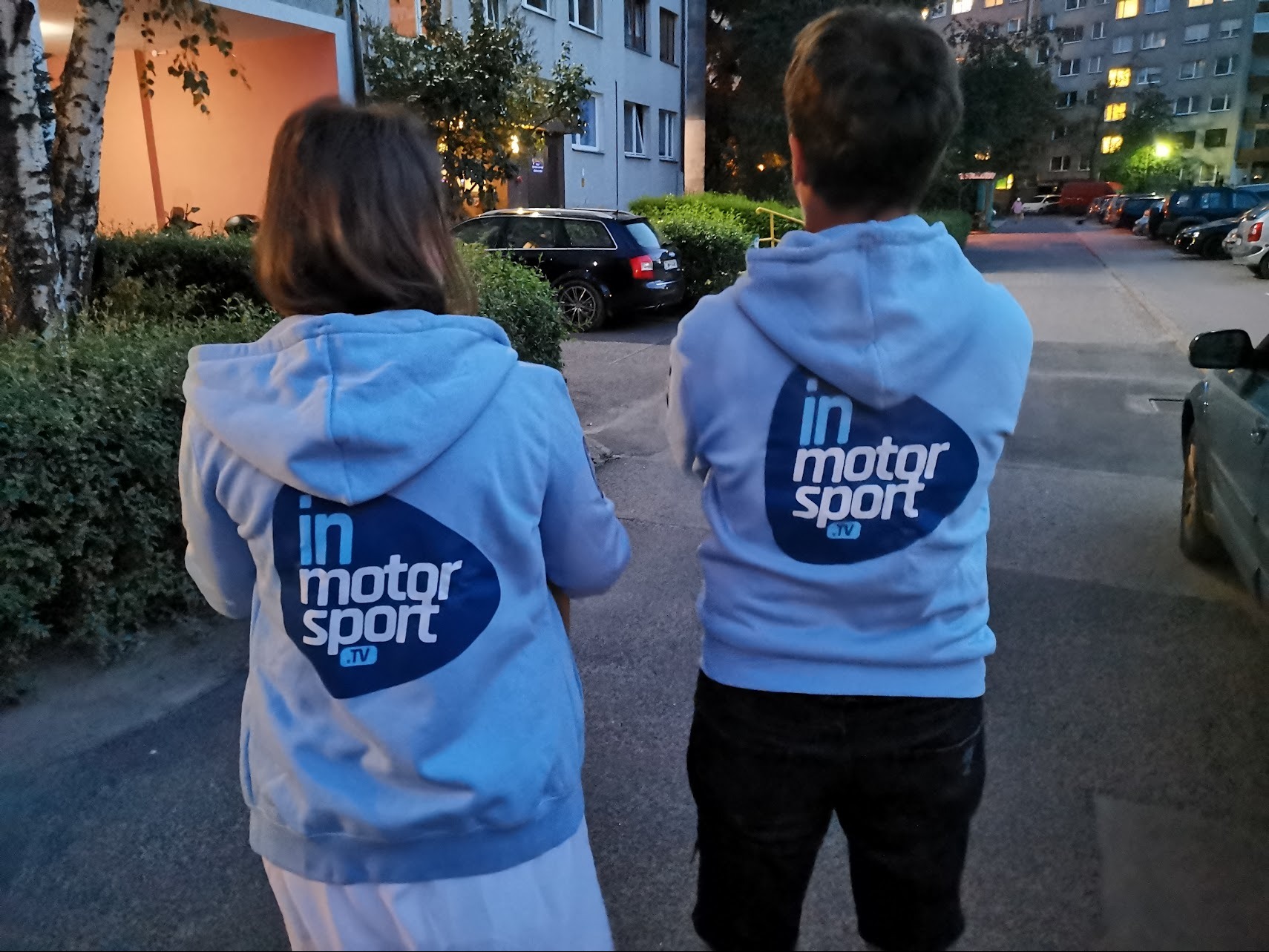 Dwie osoby stoją tyłem, ubrane w jasnoniebieskie bluzy z kapturem, na których widnieje ciemnoniebieskie logo 'in motor sport .tv', widok z tyłu na tle parkingu i bloku mieszkalnego.