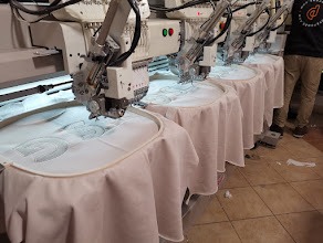 Rząd automatycznych maszyn do szycia, wykonujących identyczny wzór na białych tkaninach, naciągniętych na okrągłe tamborki. Widoczny fragment osoby obsługującej maszyny.