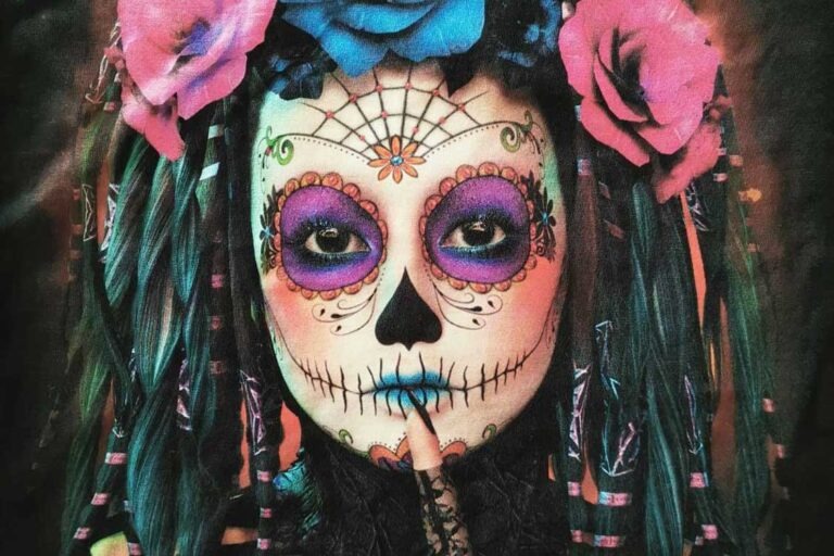Portret kobiety z makijażem w stylu meksykańskiego Dnia Zmarłych, ozdobiona kwiatami we włosach, trzymająca palec na pomalowanych na niebiesko ustach.
