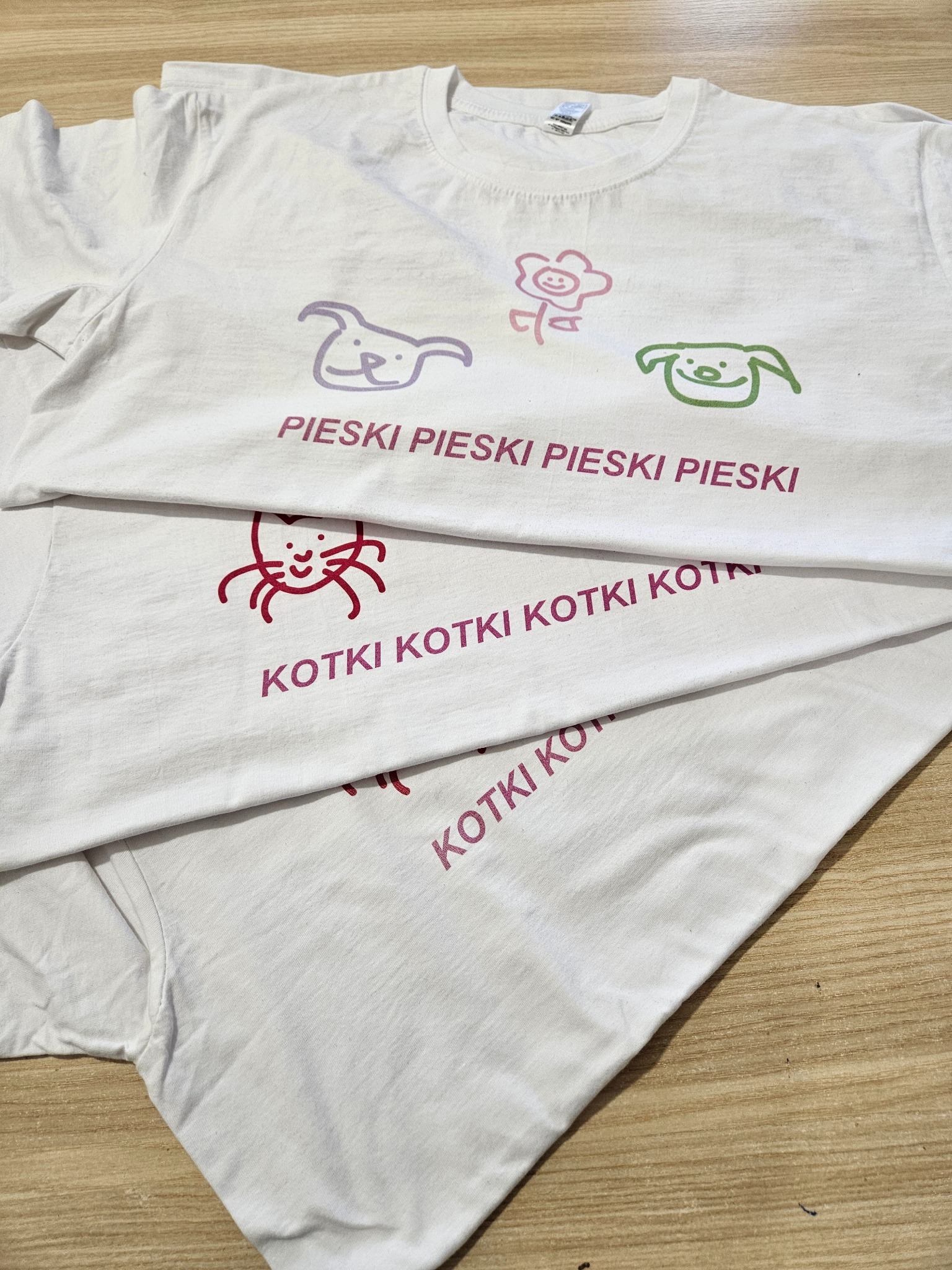 Stos białych koszulek z nadrukami: na jednej koszulce fioletowe rysunki psów i kwiatka oraz napis 'PIESKI PIESKI PIESKI PIESKI', na drugiej czerwony rysunek kota i napis 'KOTKI KOTKI KOTKI KOT'