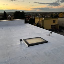 FIRMA HANDLOWO USŁUGOWA REVERSO Damian Wiecheć - Biały, płaski dach po renowacji i hydroizolacji, z widocznym świetlikiem dachowym, kominkiem wentylacyjnym i elementami instalacji odgromowej, na tle zabudowy miejskiej i zachodzącego słońca.