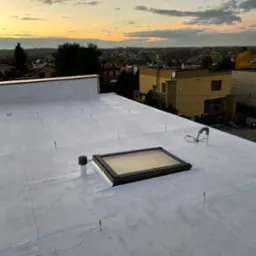 Biały, płaski dach po renowacji i hydroizolacji, z widocznym świetlikiem dachowym, kominkiem wentylacyjnym i elementami instalacji odgromowej, na tle zabudowy miejskiej i zachodzącego słońca.