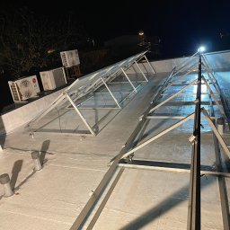 FIRMA HANDLOWO USŁUGOWA REVERSO Damian Wiecheć - Biała, hydroizolacyjna powłoka dachu z zamontowaną metalową konstrukcją pod panele fotowoltaiczne, widoczne klimatyzatory LG na krawędzi dachu oraz nocne oświetlenie.