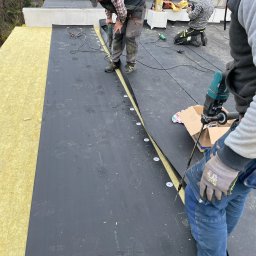FIRMA HANDLOWO USŁUGOWA REVERSO Damian Wiecheć - Pracownicy montują membranę EPDM na ocieplonym dachu płaskim, kołkując ją wzdłuż krawędzi na wełnie mineralnej, widoczne ślady butów na membranie.