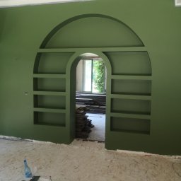 FIRMA HANDLOWO USŁUGOWA REVERSO Damian Wiecheć - Ściana w kolorze oliwkowej zieleni z wnęką w kształcie łuku i półkami, widoczne narzędzia i materiały malarskie na podłodze, w tle ułożone płyty.