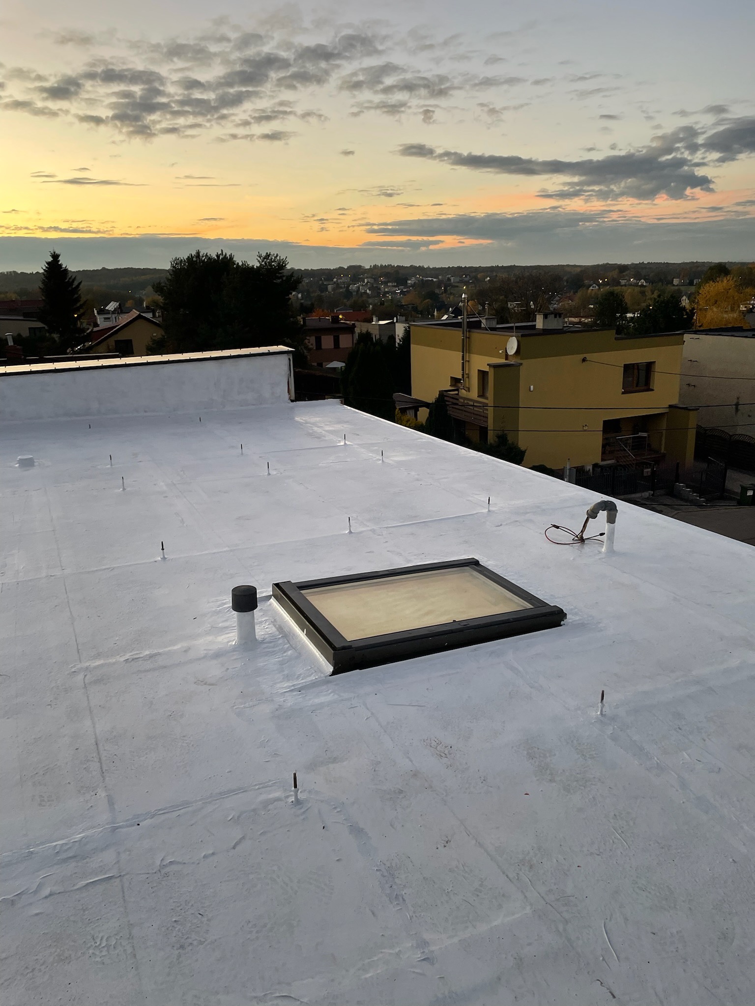 Biały, płaski dach po renowacji i hydroizolacji, z widocznym świetlikiem dachowym, kominkiem wentylacyjnym i elementami instalacji odgromowej, na tle zabudowy miejskiej i zachodzącego słońca.