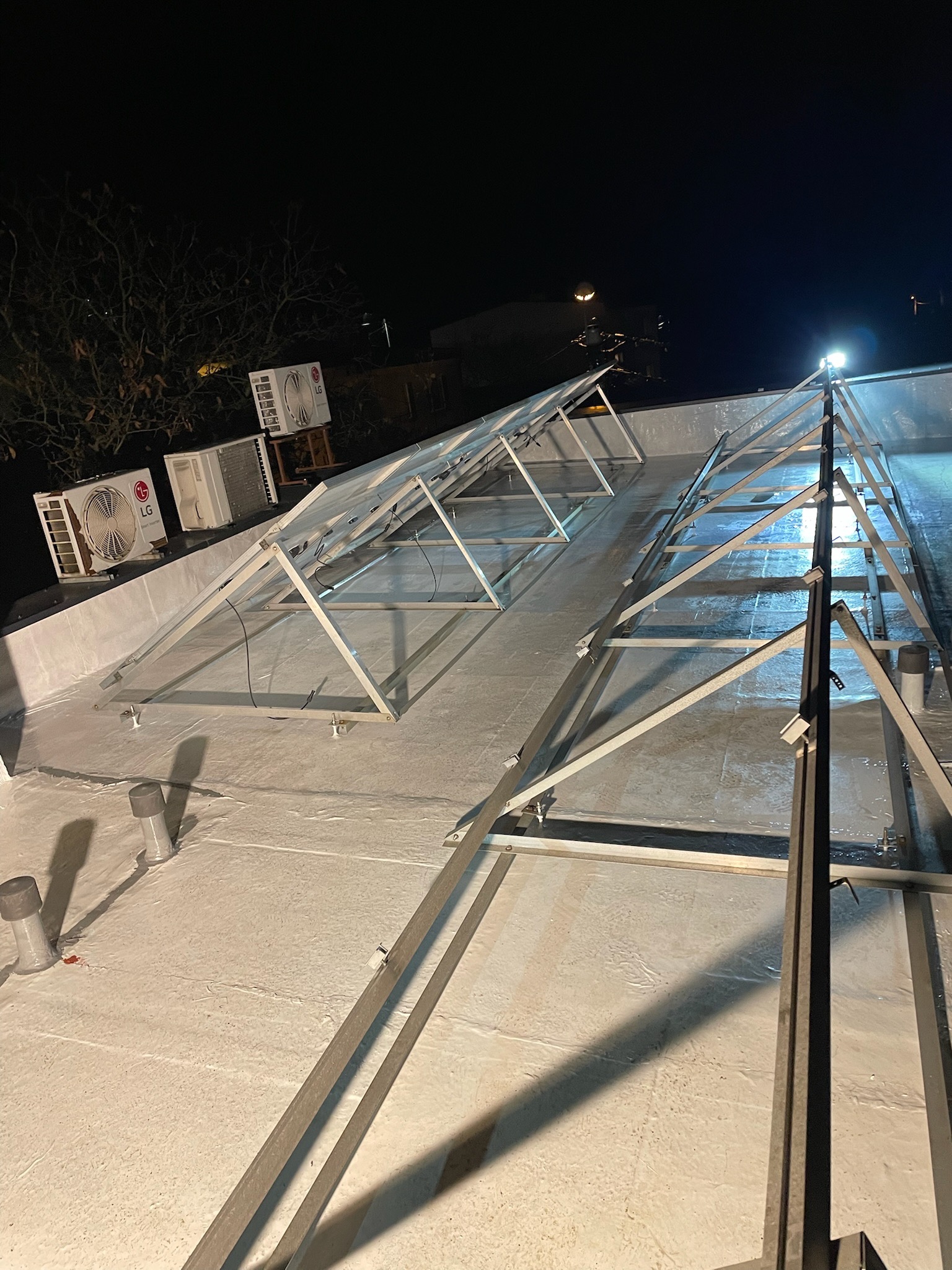 Biała, hydroizolacyjna powłoka dachu z zamontowaną metalową konstrukcją pod panele fotowoltaiczne, widoczne klimatyzatory LG na krawędzi dachu oraz nocne oświetlenie.