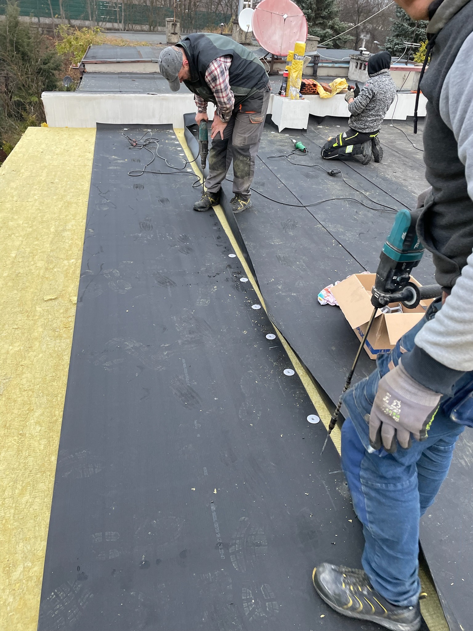 Pracownicy montują membranę EPDM na ocieplonym dachu płaskim, kołkując ją wzdłuż krawędzi na wełnie mineralnej, widoczne ślady butów na membranie.