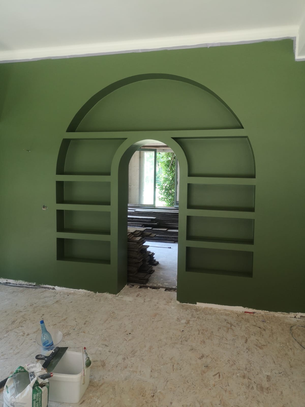 Ściana w kolorze oliwkowej zieleni z wnęką w kształcie łuku i półkami, widoczne narzędzia i materiały malarskie na podłodze, w tle ułożone płyty.