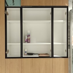 Jaqbex - Szafka łazienkowa z drewnianymi frontami, czarnymi obramowaniami i białym wnętrzem, z kosmetykami na półkach, widoczne zawiasy i fragment ściany w trakcie remontu.