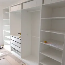 Biała, modułowa szafa z szufladami i półkami w trakcie montażu, widoczne narzędzia i plany na półce.