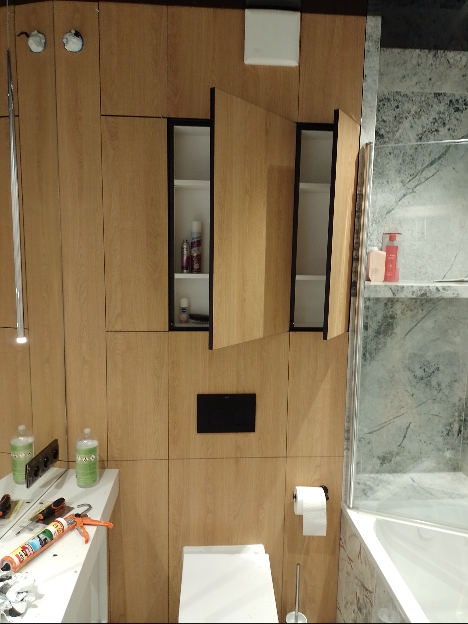 Łazienka z zabudową meblową z drewnopodobnej płyty, szafki z czarnymi obramowaniami otwarte, widoczne kosmetyki wewnątrz, na blacie silikon i narzędzia, toaleta i wanna obok.