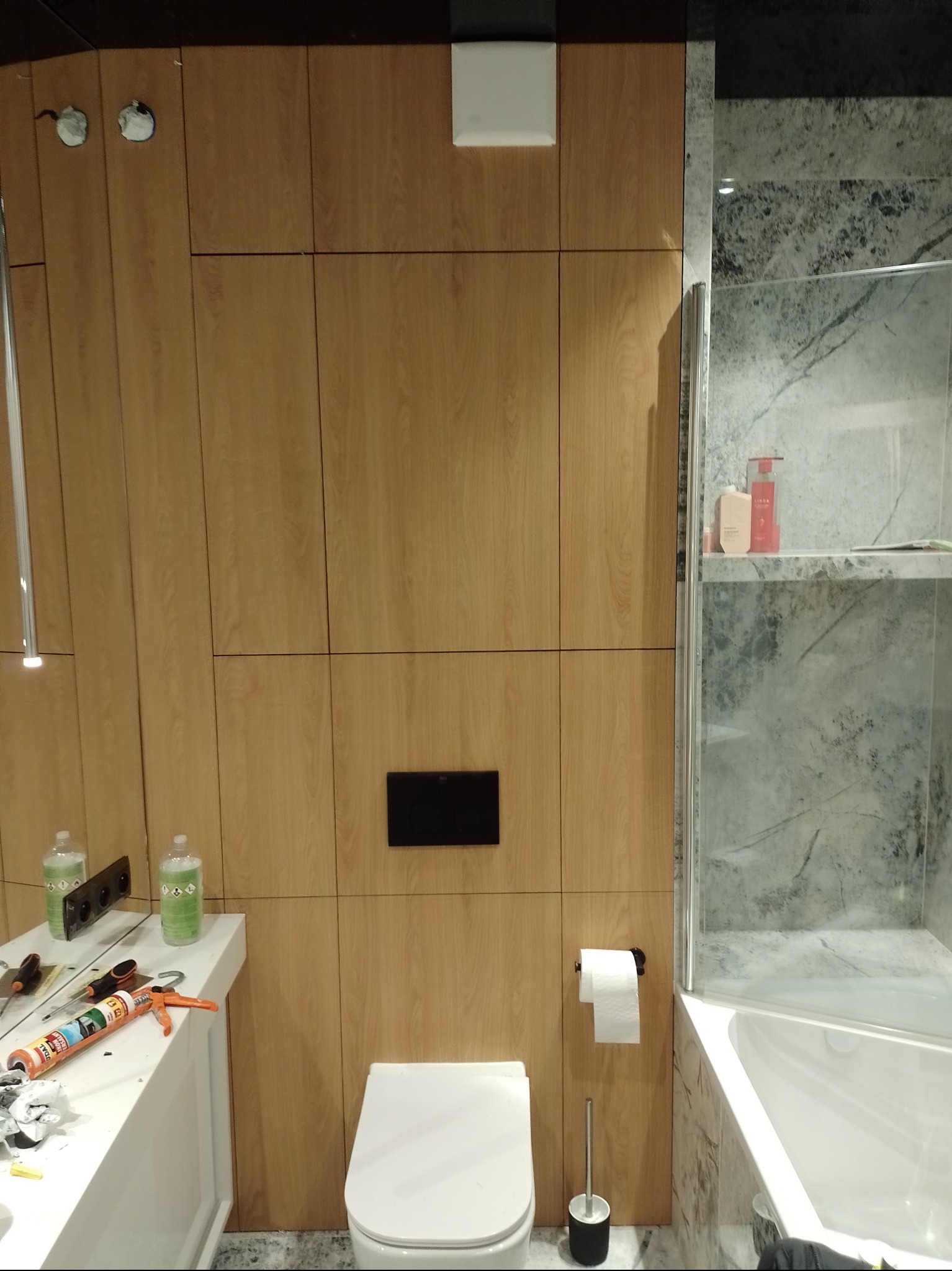 Montaż drewnianej zabudowy w łazience, widoczna toaleta, wanna z parawanem, silikon i narzędzia na blacie umywalki w trakcie pracy.