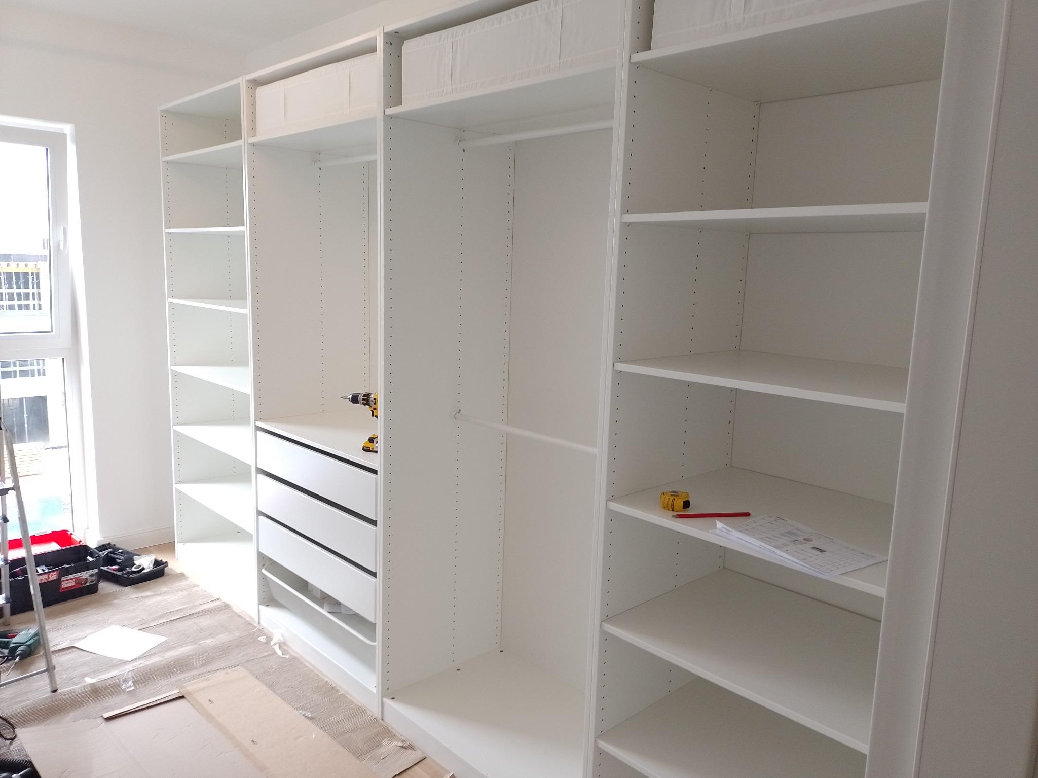 Biała, modułowa szafa z szufladami i półkami w trakcie montażu, widoczne narzędzia i plany na półce.