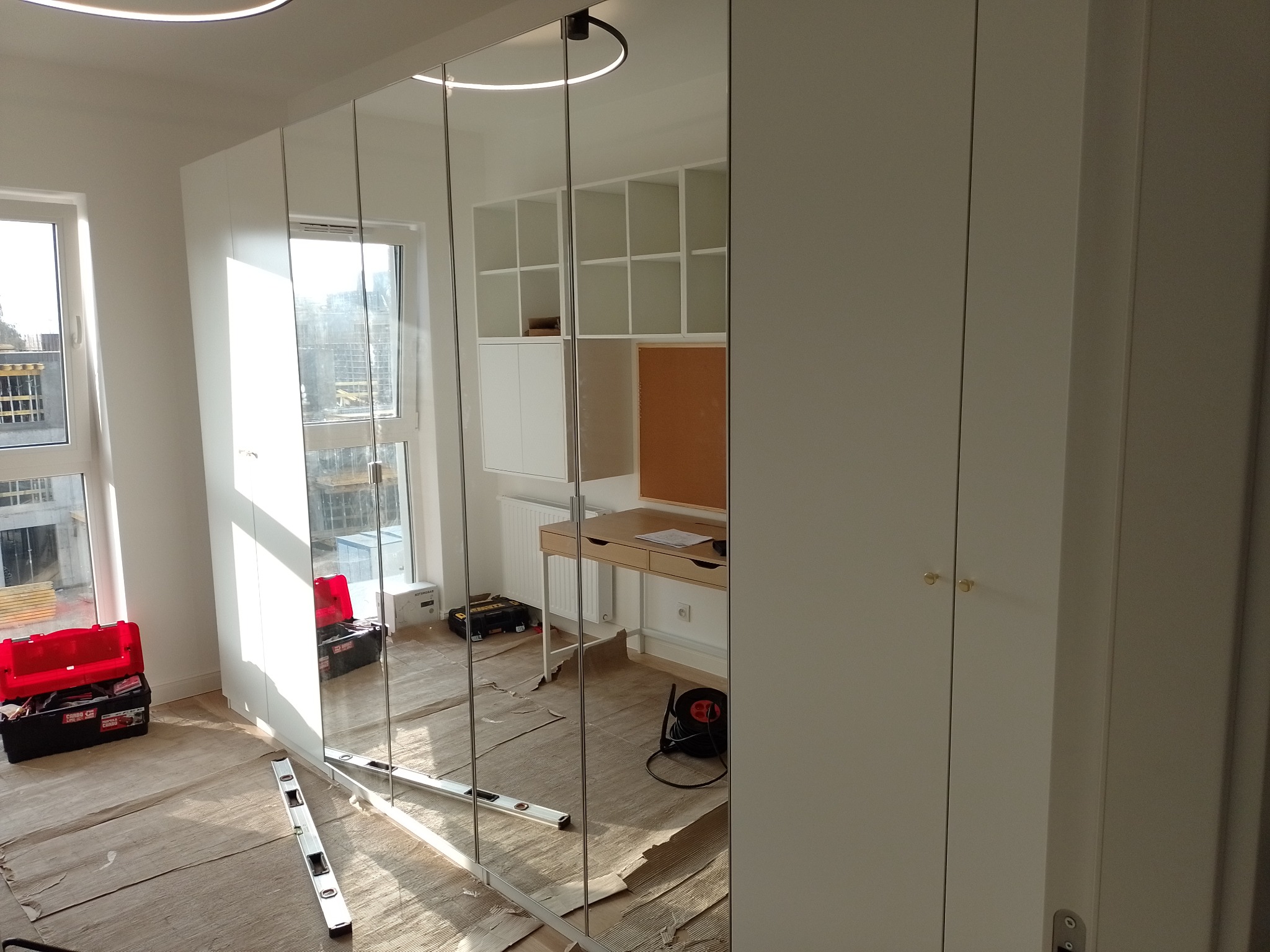 Montaż wielkogabarytowej szafy z lustrzanymi drzwiami przesuwnymi w jasnym pomieszczeniu, widoczne narzędzia i zabezpieczenia podłogi podczas instalacji.