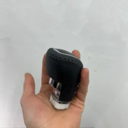 Tapicerowana, czarna gałka zmiany biegów z perforowanej skóry, trzymana w dłoni, z widocznymi szwami i chromowanymi elementami.