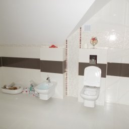 Odnowa - Biała łazienka na poddaszu z umywalką, bidetem i toaletą, wykończona płytkami w odcieniach bieli i brązu, z dekoracyjnym wzorem na ścianie i akcesoriami łazienkowymi.