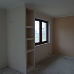 Odnowa - Pokój w trakcie wykańczania, z oknem i wnęką z półkami z płyty kartonowo-gipsowej, ściany częściowo pomalowane na beżowo, surowa podłoga, widoczne wystające kable elektryczne.