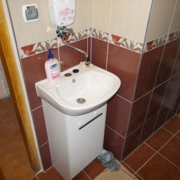 Odnowa - Mała umywalka z szafką w łazience z brązowymi płytkami, dozownik mydła i grzejnik przepływowy nad umywalką.