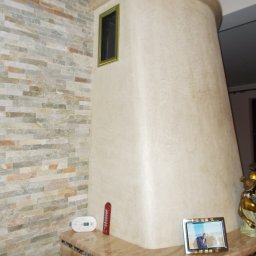 Odnowa - Obudowa kominka z beżową, teksturowaną powierzchnią, dekorowana ramką z czarną kratką, przylegająca do ściany wyłożonej kamienną mozaiką w odcieniach szarości i beżu.