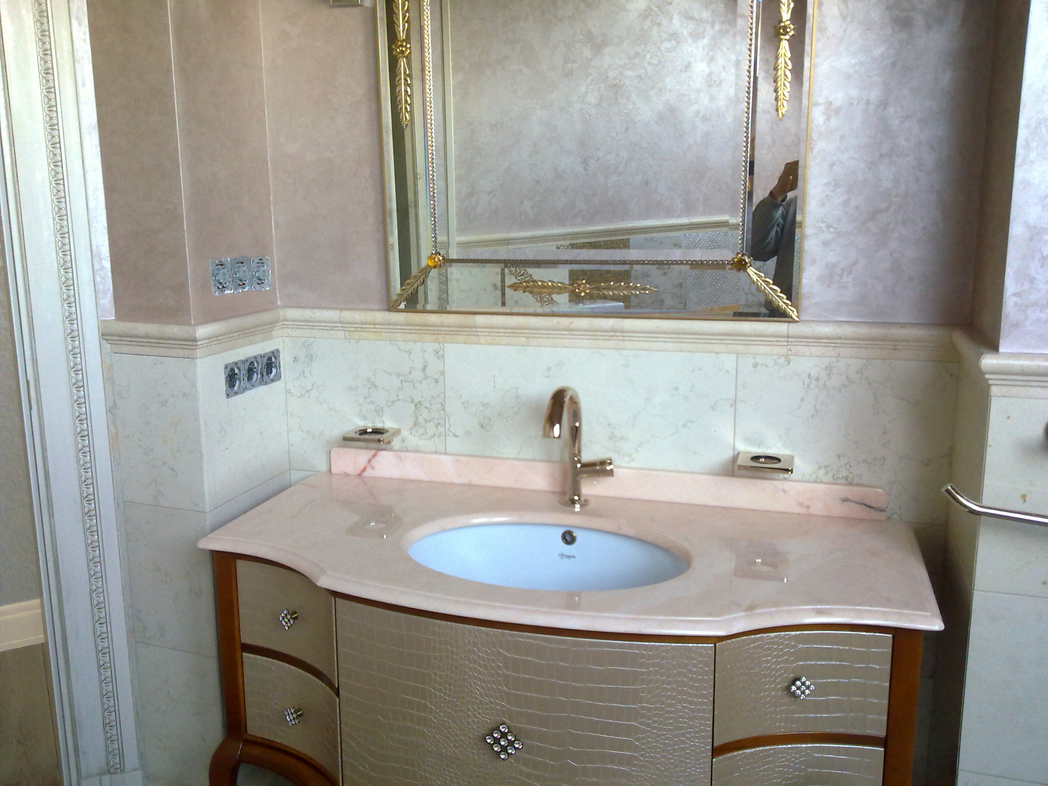 Elegancka umywalka w łazience z beżowym blatem imitującym marmur, szafką z fakturą skóry krokodyla i kryształowymi gałkami, oraz ozdobnym lustrem w złotej ramie.