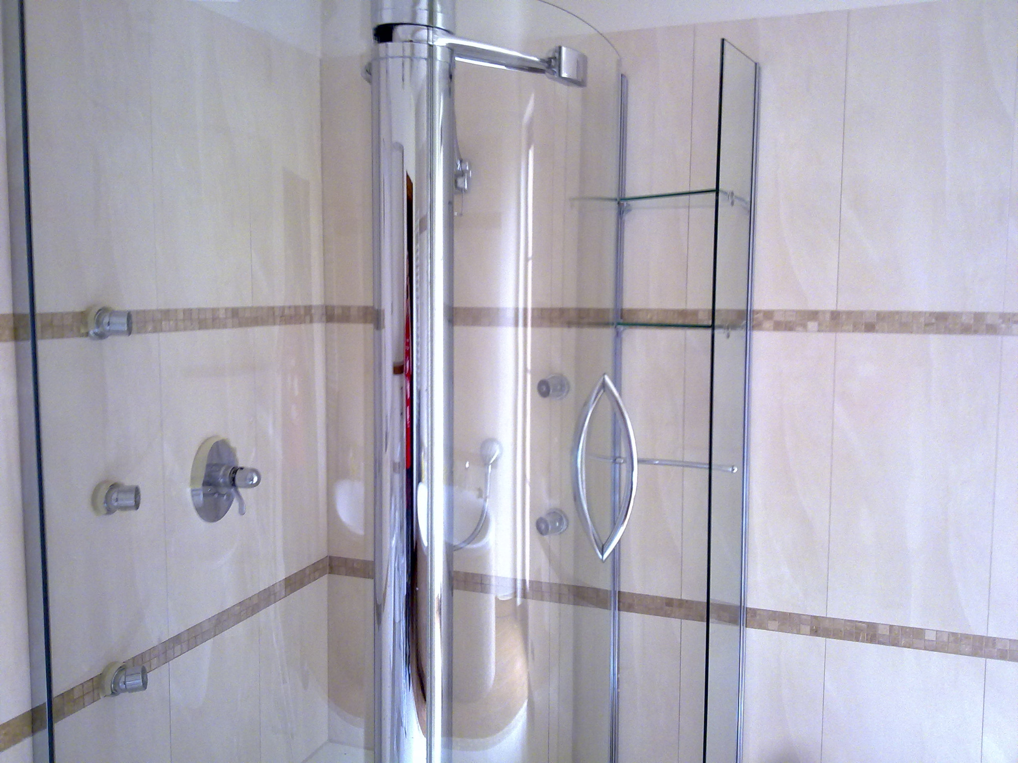 Szklana kabina prysznicowa z chromowanymi elementami, widoczne płytki w odcieniach beżu z dekoracyjnym pasem mozaiki, bateria prysznicowa zamontowana na ścianie.