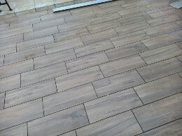 Ułożona podłoga z brązowych, prostokątnych płytek imitujących drewno, z widocznymi fugami w kolorze ciemnoszarym. Płytki ułożone w regularny wzór, z lekkim przesunięciem w każdym rzędzie. W górnej...