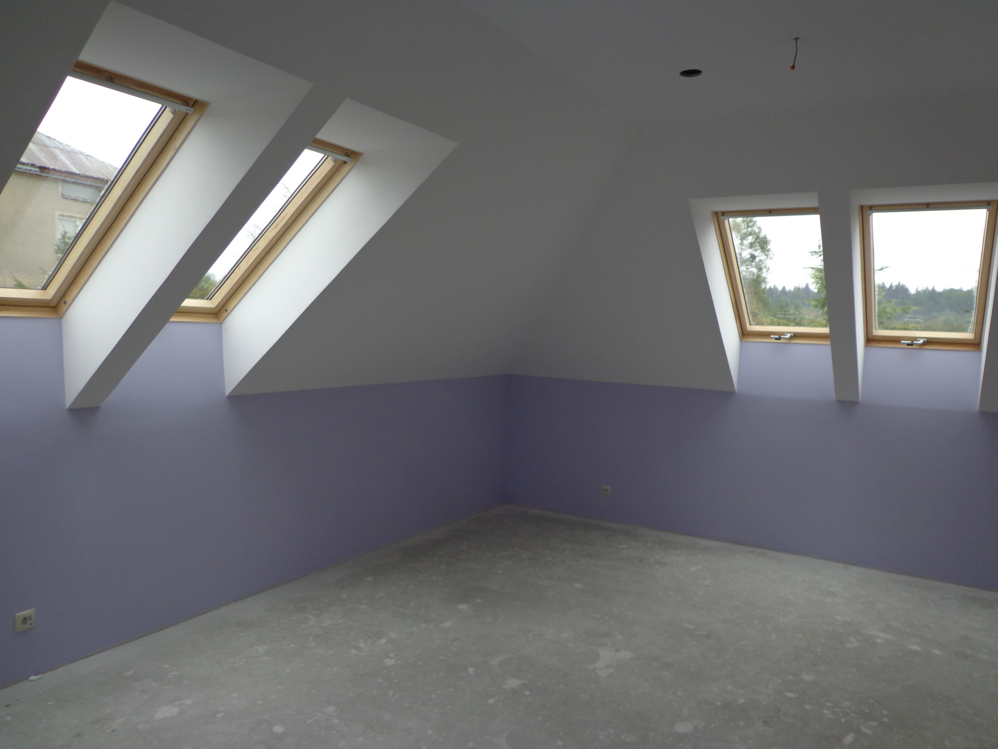 Poddasze w trakcie prac wykończeniowych: ściany pomalowane na liliowo do połowy wysokości, powyżej białe, trzy okna dachowe z drewnianymi ramami, surowa betonowa podłoga, widoczne gniazdka...