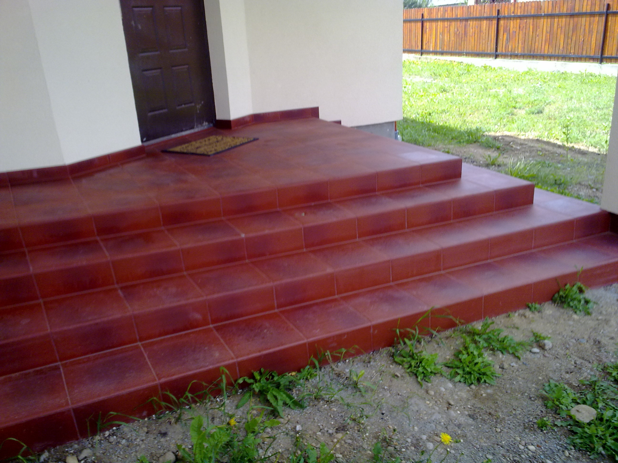 Czerwone płytki ceramiczne na schodach wejściowych do domu z brązowymi drzwiami, widok z dołu, trawa i ogrodzenie w tle.