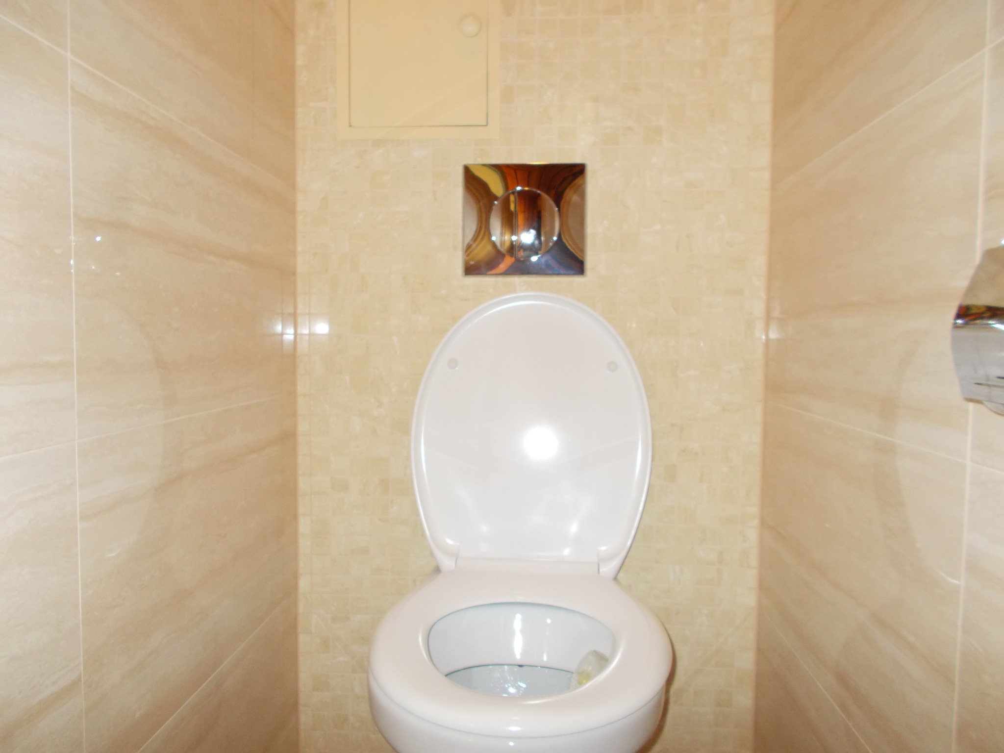 Zamontowana, biała toaleta w łazience wykończonej beżowymi płytkami, widoczny chromowany przycisk spłukiwania.